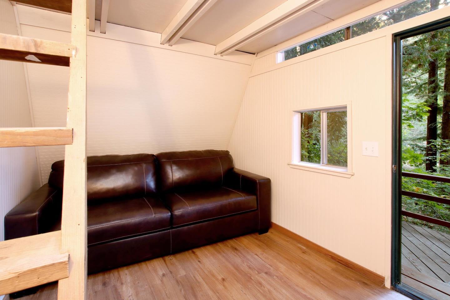 Furnished sitting area with a dark leather sofa, sloped ceiling, and glass door opening toward a wooded deck.