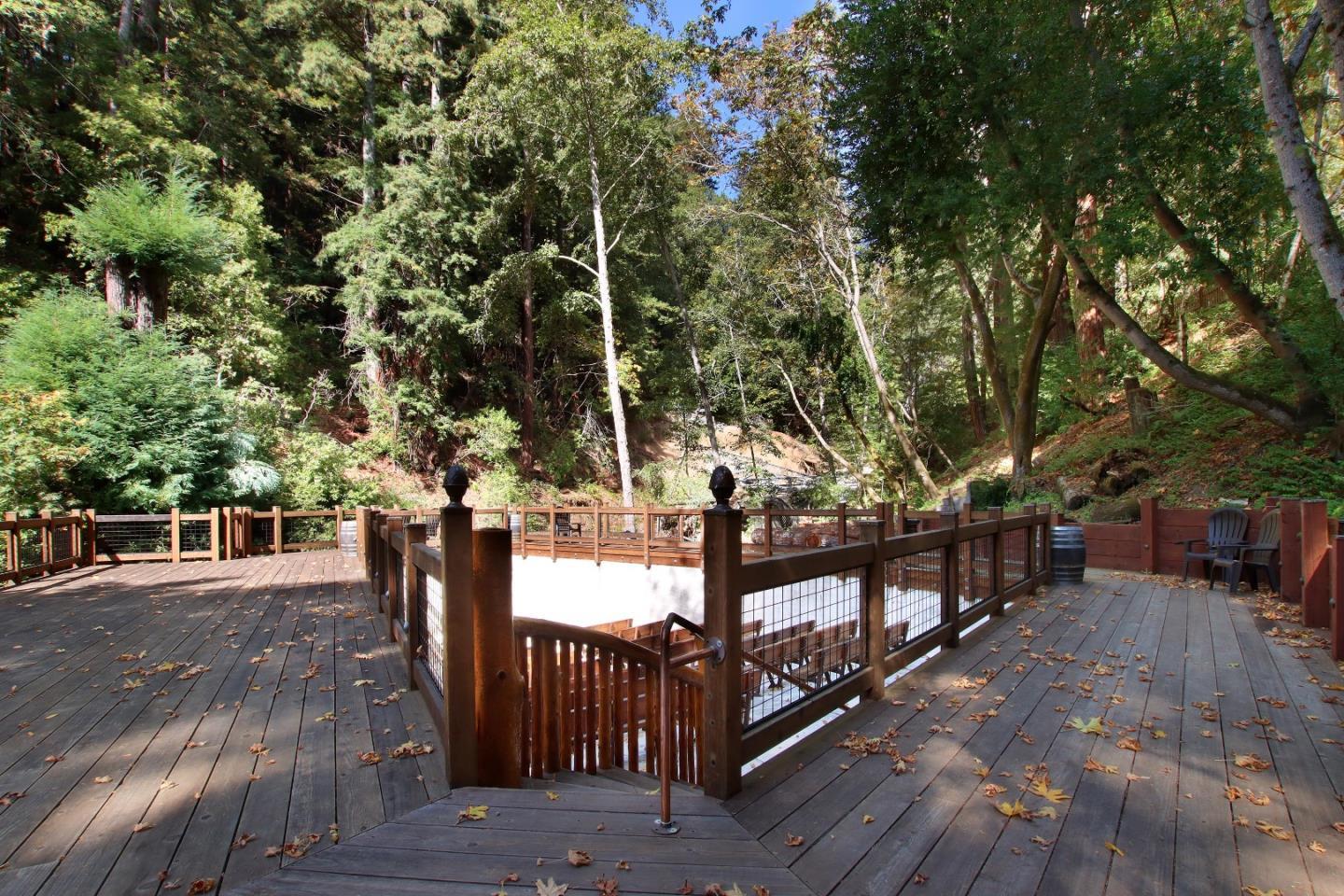 Wooden deck surrounding a swimming pool beneath dense trees, with outdoor chairs and fallen leaves.