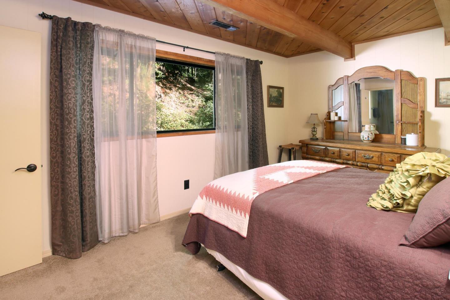 Bedroom with a large bed, wood-plank vaulted ceiling, and window overlooking a wooded hillside.
