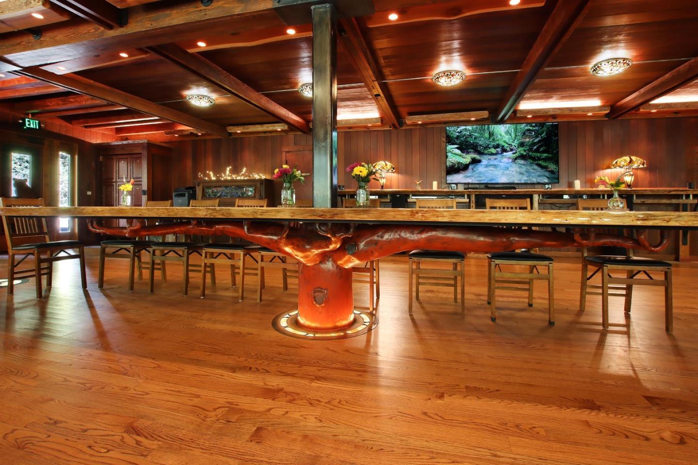 Wood-paneled gathering room with a large live-edge table, stools, and exposed ceiling beams.