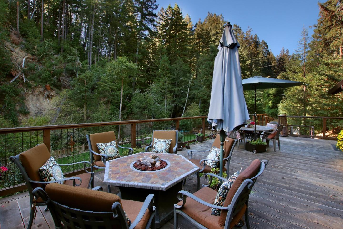 Wooded hillside deck with a tiled fire-pit table, cushioned chairs, and umbrella-covered dining area.
