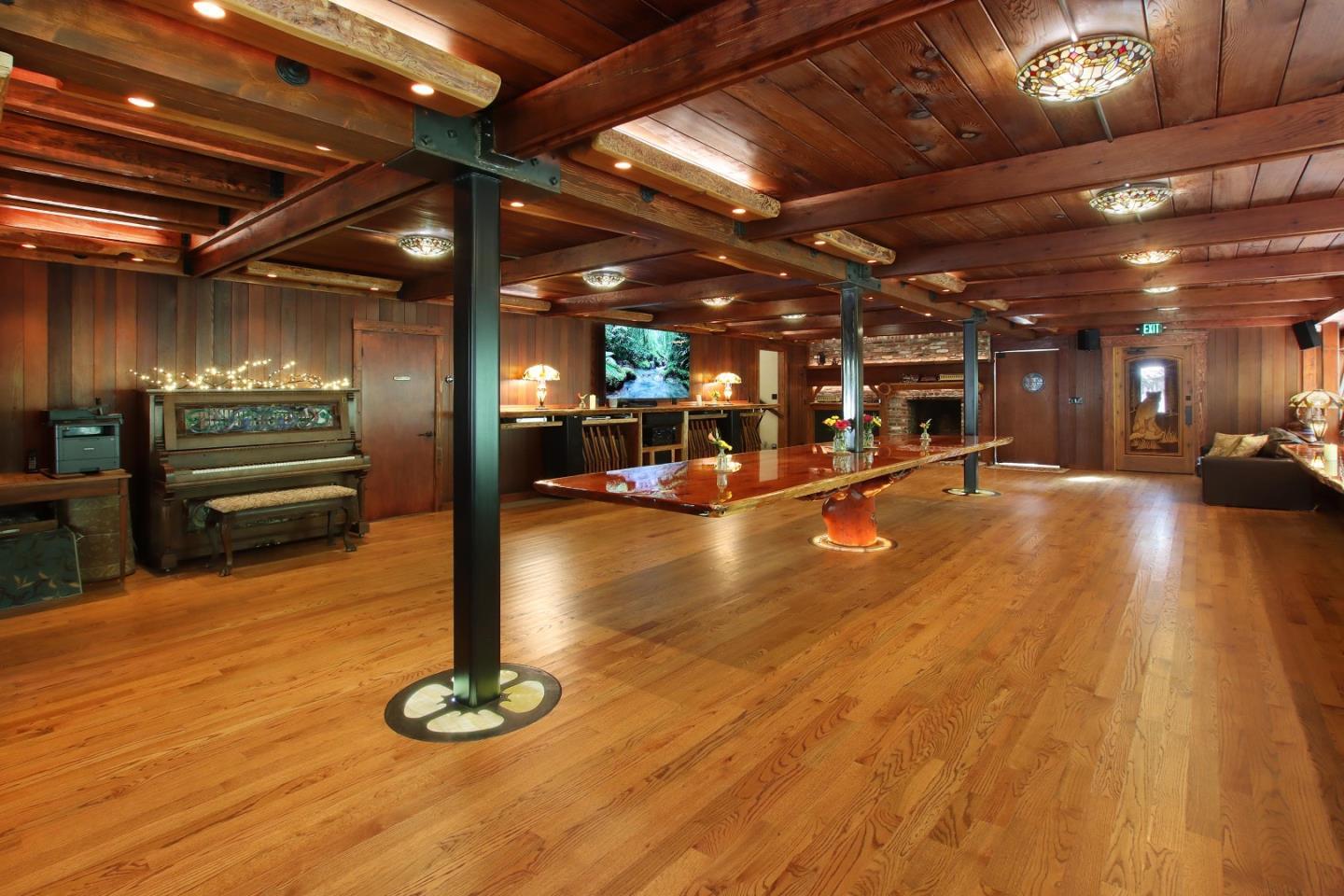 Wood-paneled great room with exposed beams, a polished table, piano, and stone fireplace.