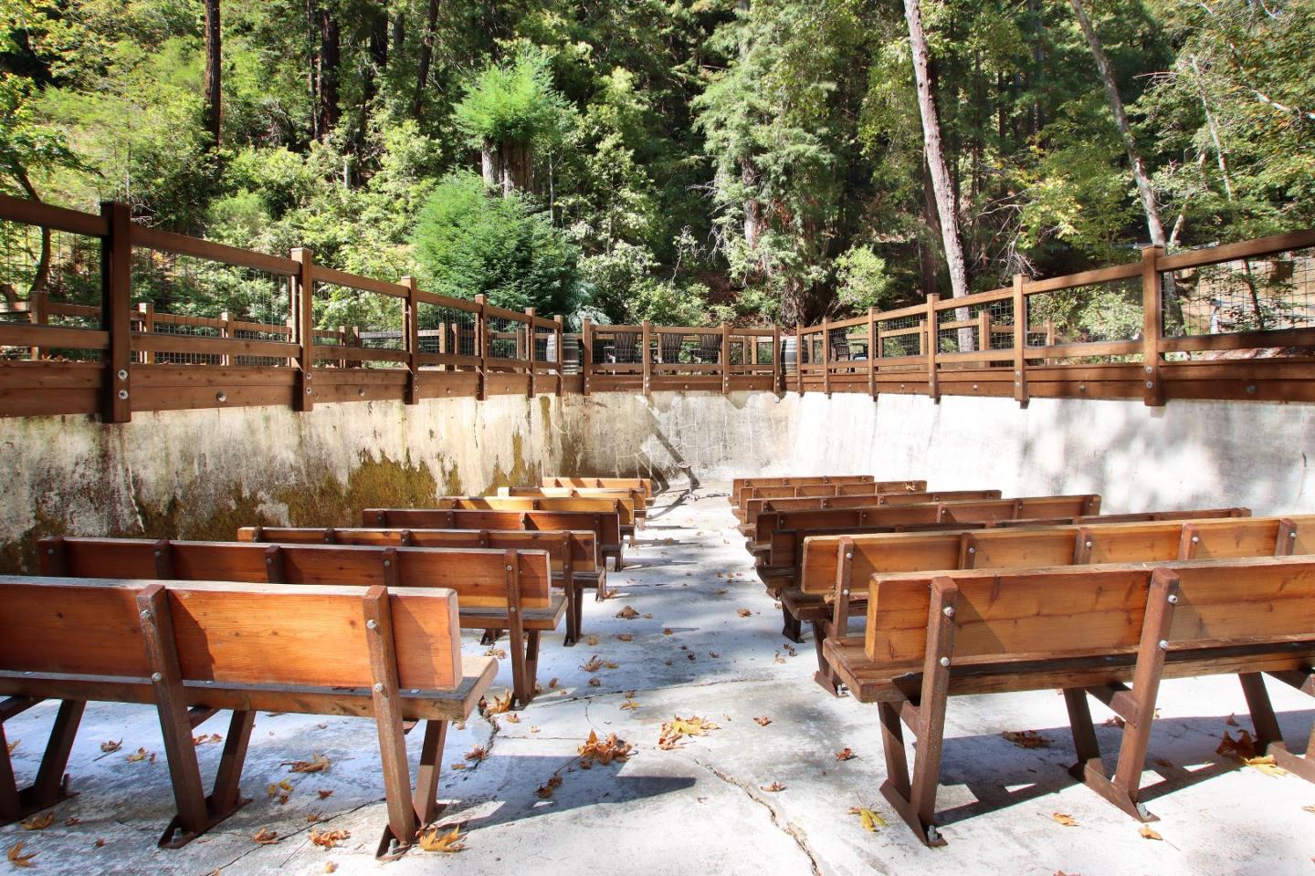 Outdoor amphitheater with rows of wooden benches surrounded by dense trees.